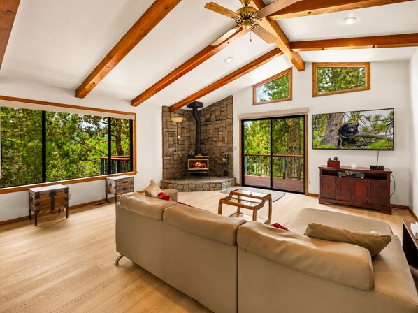 A spacious living room with a stone fireplace, large windows, and a ceiling fan. - Silver Rays - Walk to Lake/Village, Game Room, Pool Table, Air Hockey, BBQ (Big Bear Lake)