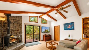 A spacious living room with a stone fireplace, wooden beams, a ceiling fan, and a large window overlooking a forest.