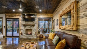 A spacious living room with a stone fireplace, leather sofa, and a large TV mounted on the wall. - Creekside 6BR Retreat w/ UTV Trails & Hot Tub & Pet-Friendly (Broken Bow)