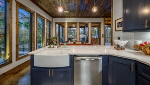 A modern kitchen with a large island, wooden ceiling, and large windows offering views of a deck and trees. - Creekside 6BR Retreat w/ UTV Trails & Hot Tub & Pet-Friendly (Broken Bow)