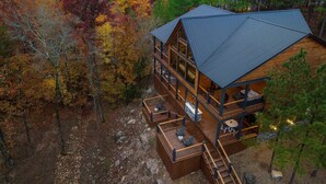 A cabin with a large deck and multiple windows, surrounded by autumn-colored trees. - Creekside 6BR Retreat w/ UTV Trails & Hot Tub & Pet-Friendly (Broken Bow)