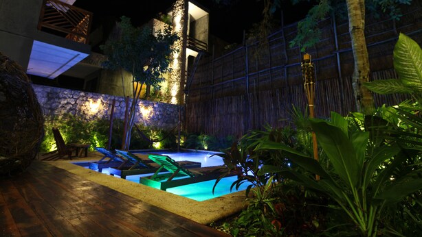A nighttime view of a swimming pool area with a wooden deck, stone wall, and tropical plants. - One-bedroom retreat with a private pool patio: your oasis in Tulum! (Tulum)