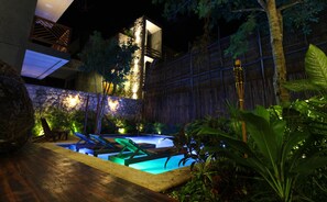 Ein Nachtblick auf einen Poolbereich mit Holzterrasse, Steinmauer und tropischer Bepflanzung.