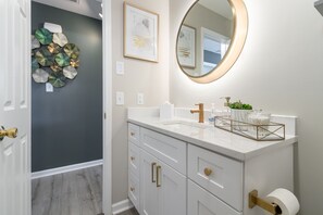 A bathroom with a white vanity, gold handles, a round mirror, and a decorative wall piece. - Graham Getaway Near Elon University (Graham)