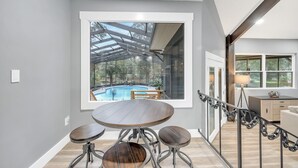 A modern interior with a large window overlooking a swimming pool, a round dining table with stools, and a staircase with a metal railing. - Beautiful Lake getaway with pool, upgraded, pet friendly with lots of extras!! (Green Cove Springs)