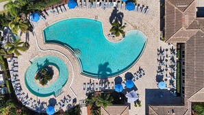 An aerial view of a resort with a large central pool, smaller circular pools, and numerous sun loungers and umbrellas. - Luxury Condo at River Strand Golf & Country Club - 2027 Available! (Bradenton)