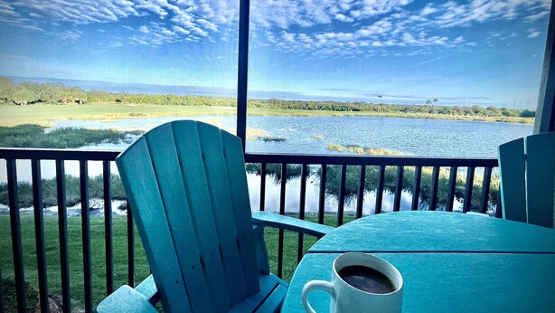 A view from a balcony with a table and chairs, overlooking a body of water and a golf course. - Luxury Condo at River Strand Golf & Country Club - 2027 Available! (Bradenton)