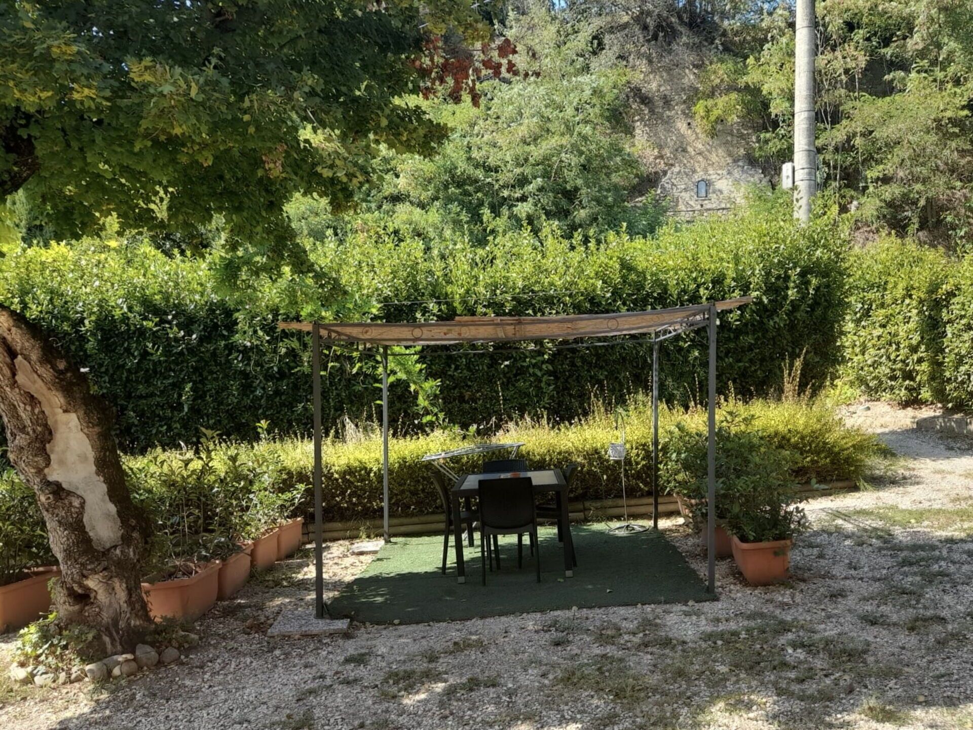 Un área de estar al aire libre cubierta, con una mesa y sillas, rodeada de plantas en macetas y un camino de grava.