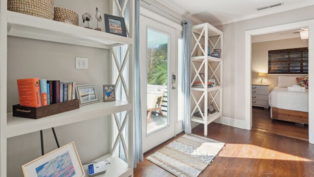A well-lit room with a wooden floor, a bed with white bedding, a desk with a computer, and shelves with books and decorative items.