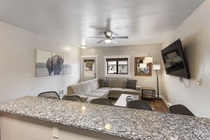 A modern living room with a large sectional sofa, a granite countertop, and a flat-screen TV mounted on the wall.