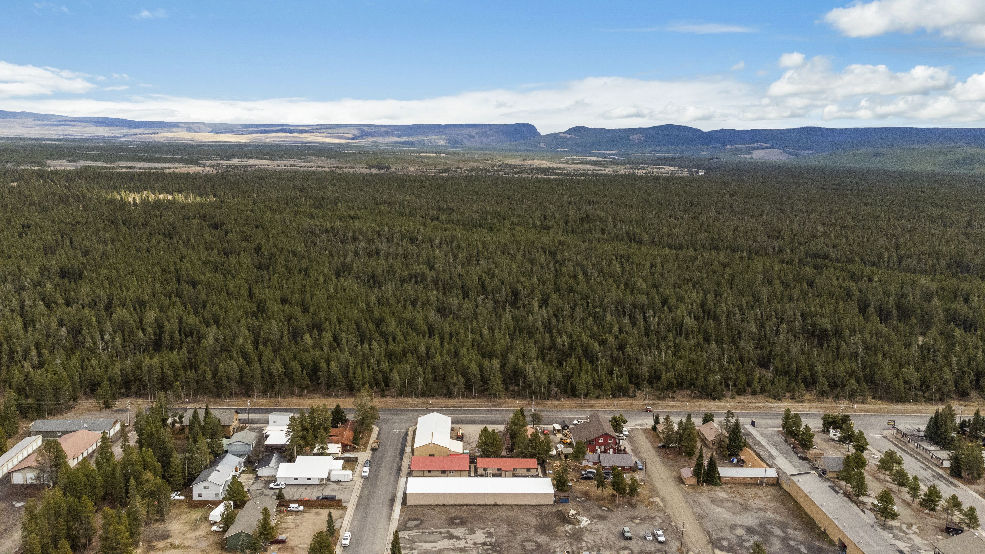 An aerial view of a small town with dense forest in the background, a main road, and several buildings including a large warehouse and residential houses.