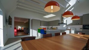 Cuina moderna amb una illa gran, llums penjants i zona de dinar amb taula i cadires de fusta.