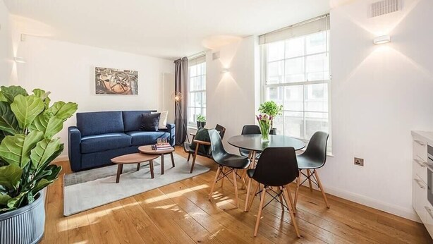 A modern living room with a blue sofa, a dining table with chairs, and a large window providing natural light. - Fantastic 1 Bedroom Flat Leicester Square West End (Greater London)