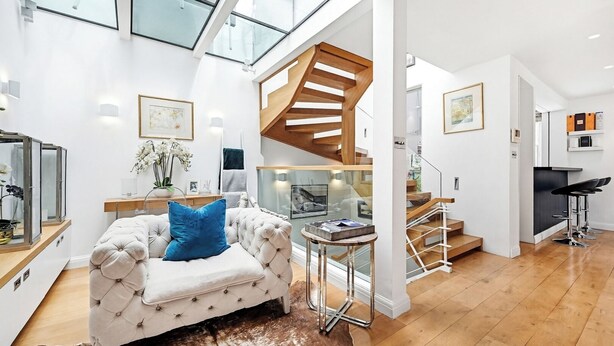 A modern interior with a tufted sofa, a glass coffee table, a wooden staircase, and a kitchen area.