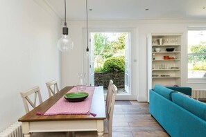A dining area with a wooden table, chairs, and a pink placemat. There is a teal sofa and a shelving unit with glassware.