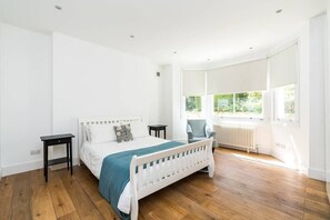 A bedroom with a white bed, a blue throw, a white radiator, and a window with blinds.