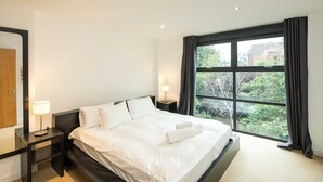 Une chambre d’hôtel moderne avec un grand lit, deux tables de chevet, une petite table, une fenêtre avec des rideaux et une vue sur la verdure environnante.