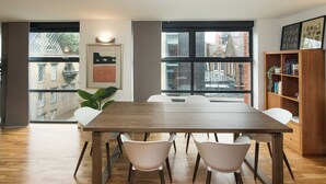 Un espace repas moderne avec une table en bois et des chaises blanches, de grandes fenêtres offrant une vue sur les bâtiments, et une bibliothèque contenant des livres et des objets décoratifs.