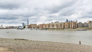 A river with buildings along the banks, a person standing on the shore, and a cloudy sky.