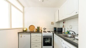Una cocina compacta con muebles blancos, horno, cocina y lavavajillas. Hay una ventana a la izquierda y un fregadero a la derecha.