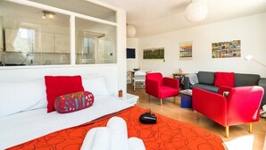 Una habitación moderna con una cama, un sofá, un sillón rojo y un sofá gris. Hay una ventana, una mesa y una pared con cuadros.