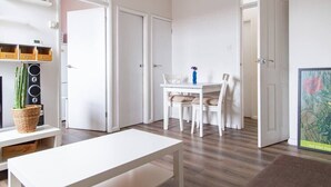 Een moderne, minimalistische woonkamer met een witte tafel, een witte bank en een eethoek met een witte tafel en stoelen. Er zijn witte deuren die leiden naar andere kamers, en een ingelijst kunstwerk op de muur.