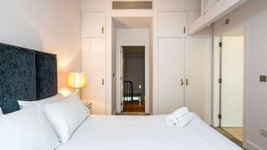 A hotel room with a bed, a nightstand, a lamp, a wardrobe, and a door leading to a hallway. - A Stunning Three Bedroom Home in Mayfair (Greater London)