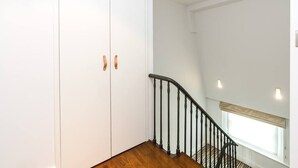 A staircase with a wooden floor and black balusters, leading to a room with white walls and a window with a beige blind. - A Stunning Three Bedroom Home in Mayfair (Greater London)