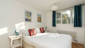 Ein Schlafzimmer mit einem großen Bett, weißen Wänden und zwei Postern. Es gibt ein Fenster mit Vorhängen, einen Nachttisch mit einer Lampe und eine Heizung.