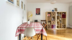 Ein Essbereich mit einem Tisch, der mit einer karierten Tischdecke bedeckt ist, einem Stuhl, einer Topfpflanze und einem Bücherregal mit verschiedenen Gegenständen.