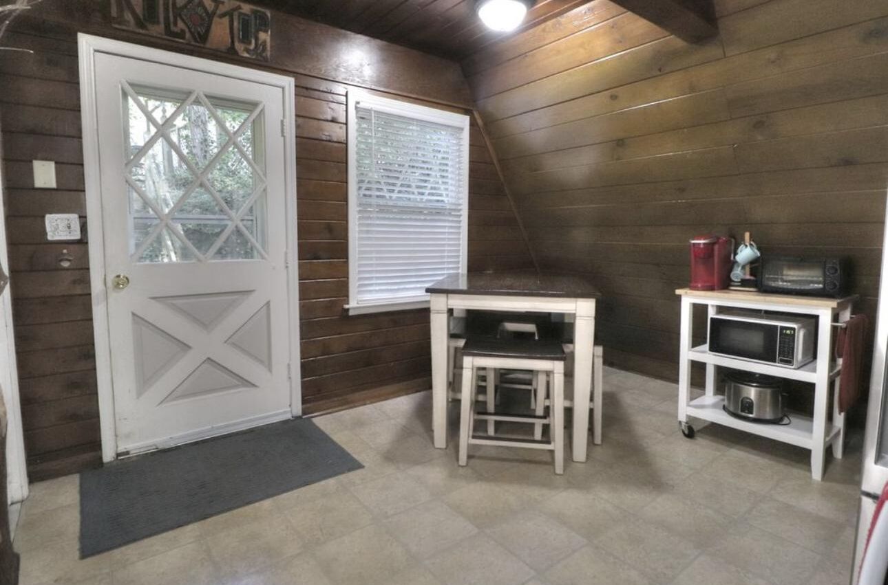 A small, cozy kitchen with wooden walls, a white door, a window with blinds, a small table, and a microwave on a cart.