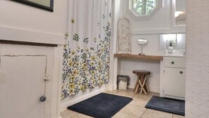 Un pasillo estrecho con suelo de madera, una puerta blanca, un cuadro floral enmarco, una cortina con motivos florales, un taburete pequeño, un espejo y una ventana con vistas a árboles.