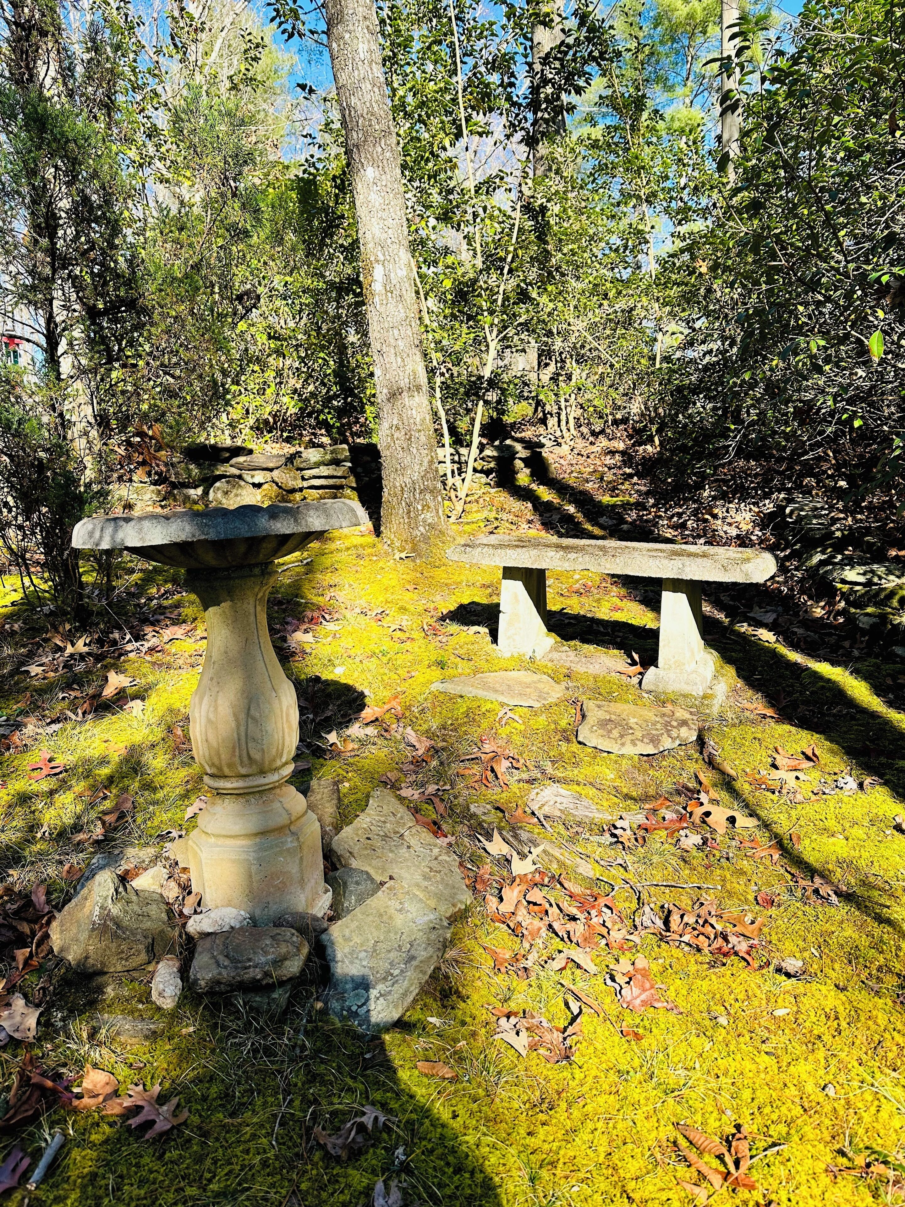 A stone bench and a stone table in a forested area with moss and fallen leaves.