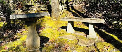 A stone bench and a stone table in a forested area with moss and fallen leaves.