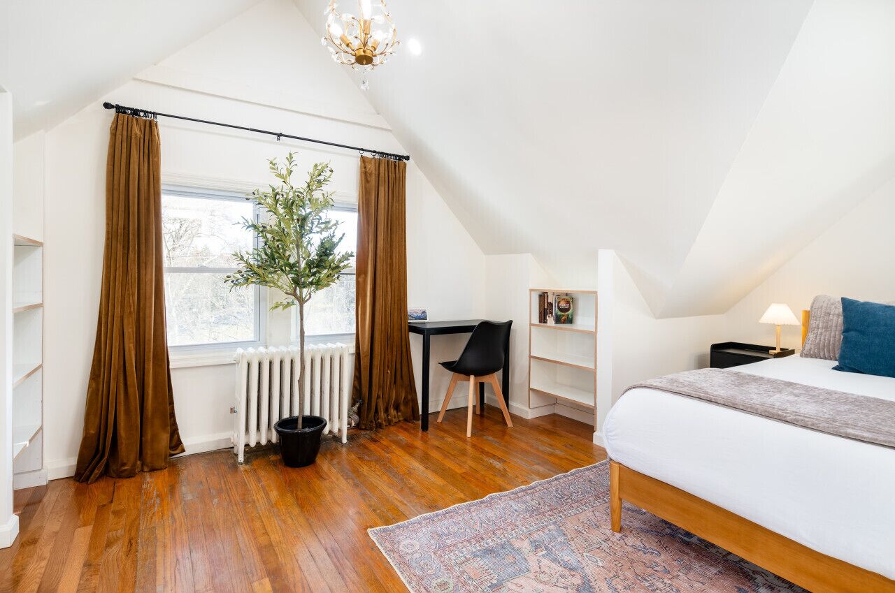 A bedroom with a sloped ceiling, a bed, a desk, a chair, a radiator, and a window with curtains.