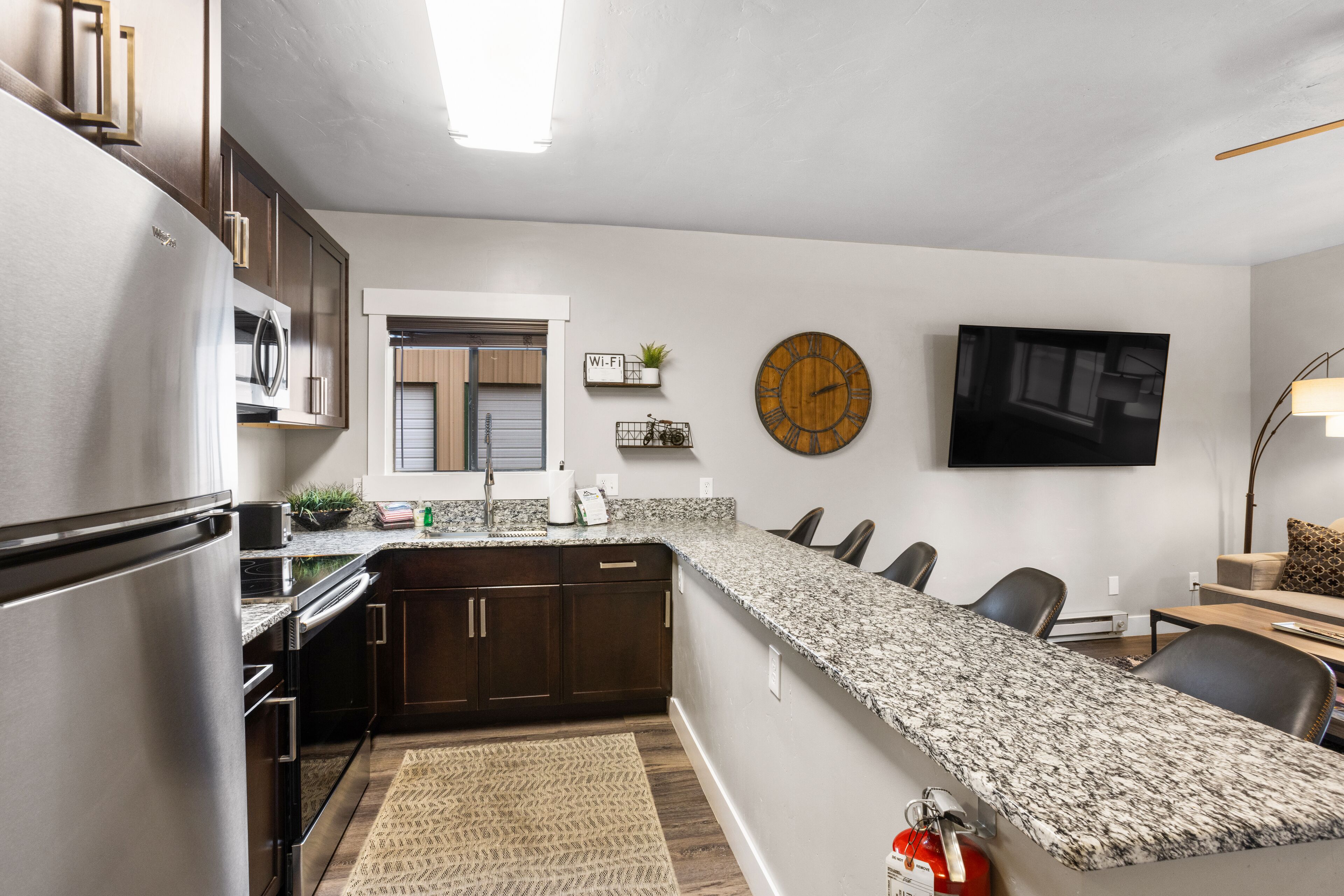 A modern kitchen with dark wood cabinets, a granite countertop, a built-in oven, and a stainless steel refrigerator.