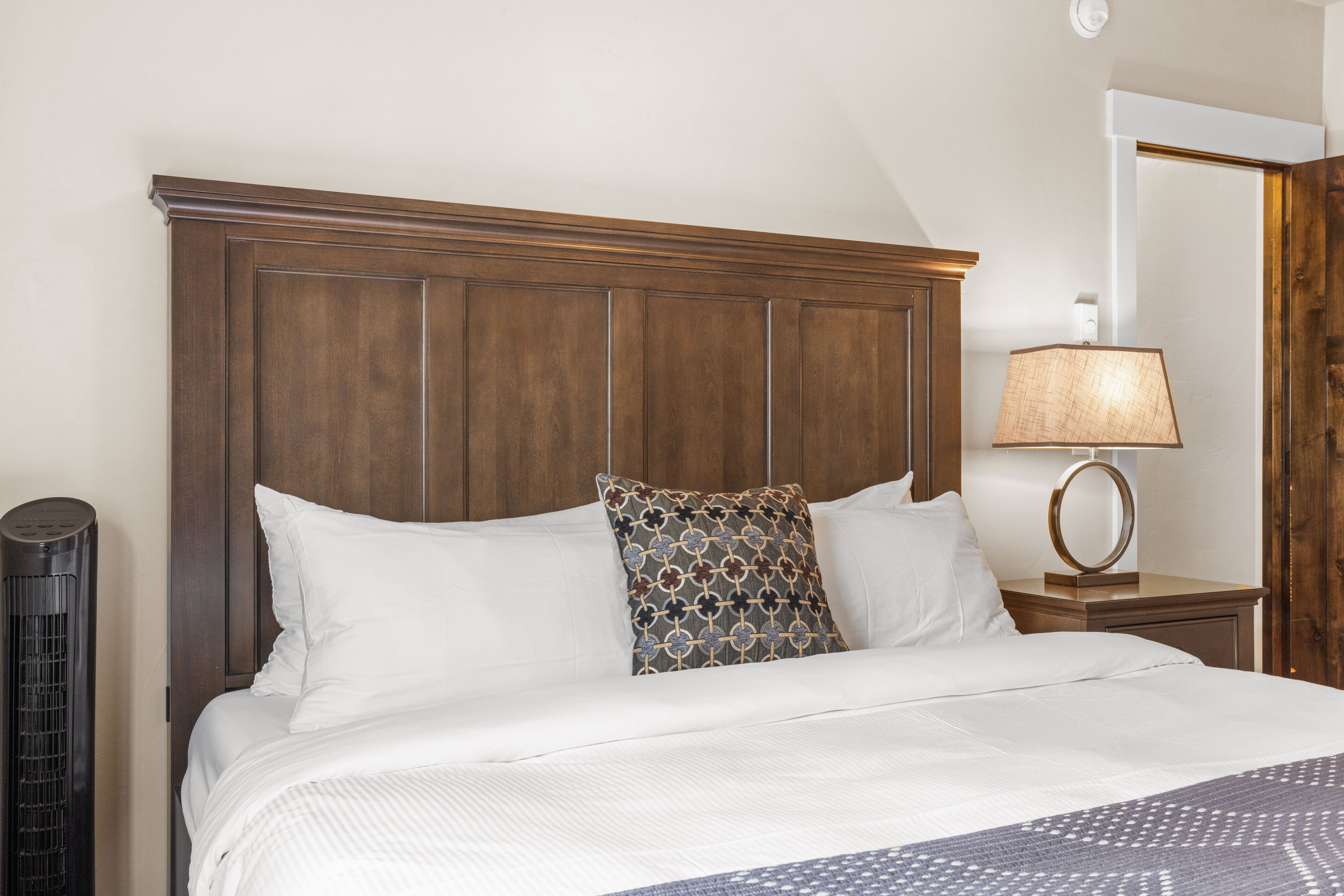 Una cama bien hecha con sábanas blancas y dos almohadones decorativos, una lámpara de noche y una lámpara de pie alta.