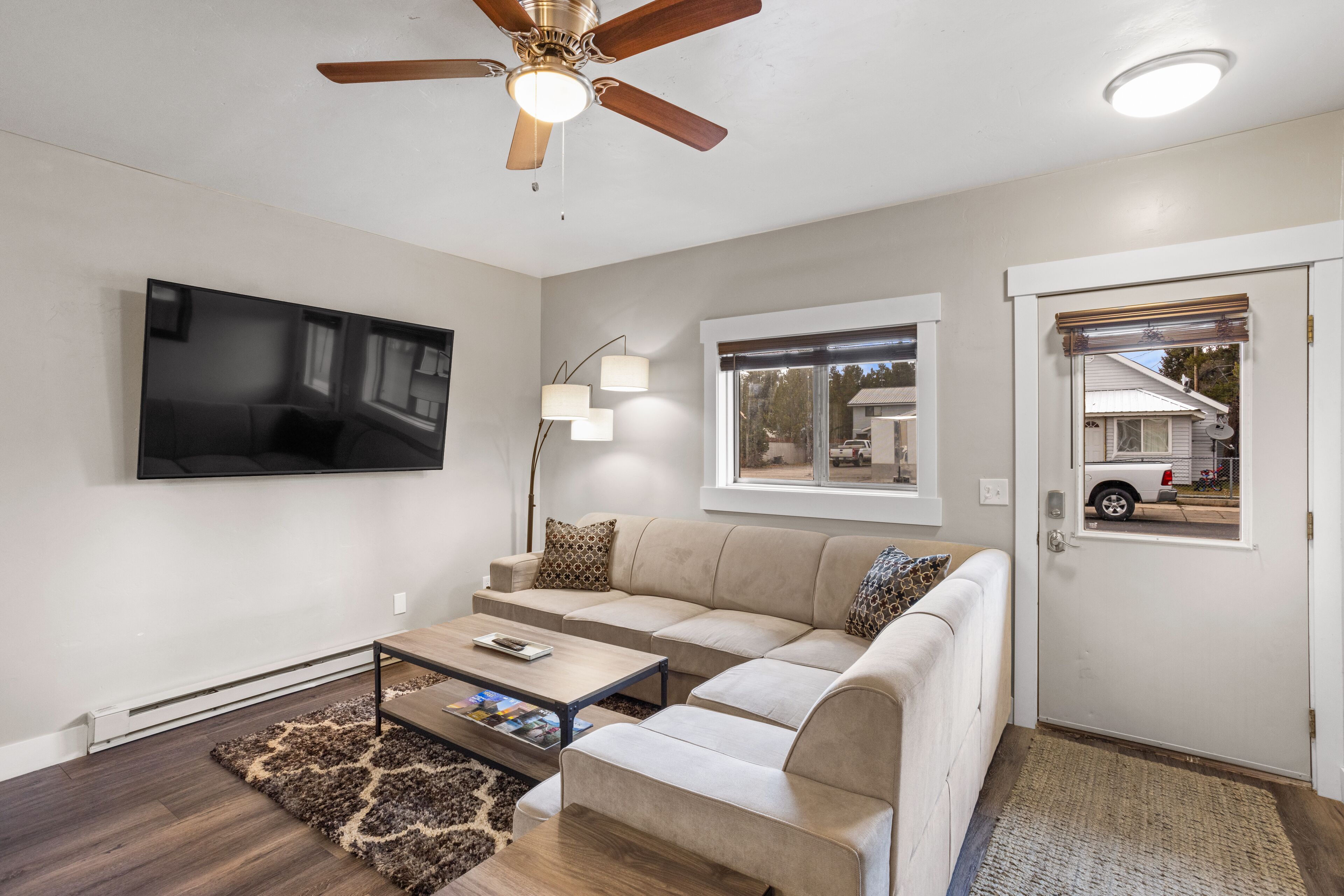 A living room with a beige sofa, a coffee table, a TV mounted on the wall, a ceiling fan, and a window with a view of a house outside.