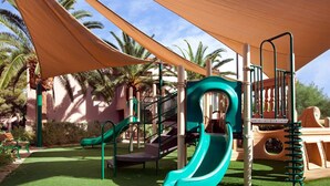 Un’area giochi con scivoli e strutture per arrampicarsi, circondata da palme e una zona all’ombra.