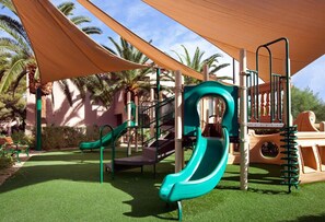Ein Spielplatz mit Rutschen und Klettergerüsten, umgeben von Palmen und einem schattigen Bereich.