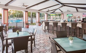 Et udendørs barområde med borde og stole, en pool i baggrunden og en bar med forskellige flasker.