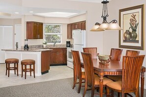 A kitchen with wooden cabinets, a central island, bar stools, a dining table with chairs, and a hanging light fixture.