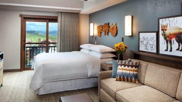 Habitación de hotel con una cama grande, un sofá, una mesa de centro con frutas y libros, un balcón con vistas y obras de arte en la pared.