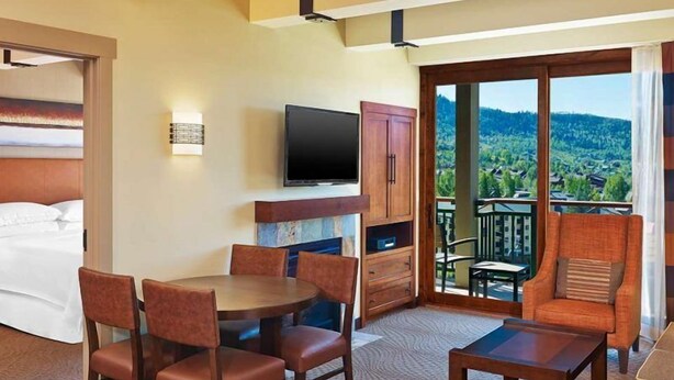 Habitación de hotel con cama, televisión, zona de comedor con sillas, mesa de centro y balcón con vistas al paisaje.