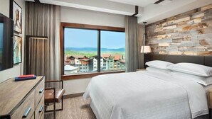 Una camera d'albergo con un letto grande, una scrivania con cassetti, una sedia, una TV e una finestra con vista su edifici e colline.