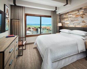 A hotel room with a large bed, a desk with drawers, a chair, a TV, and a window with a view of buildings and hills. - Steamboat Springs | Mountain Ski Lodge | Ski Access & Hot Tub | 2BR (Steamboat Springs)