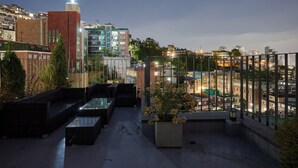 Una terraza en la azotea con mobiliario exterior, una jardinera grande con flores y vistas panorámicas de la ciudad.