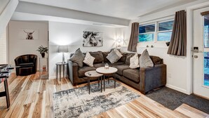 A modern living room with a brown leather sofa, a small round coffee table, and a patterned rug. There is a lamp on a side table, a wall-mounted artwork, and a window with curtains. - The Apres Chalet on Peak 7, Private Hot Tub, Locaton & Great Views! (Breckenridge)