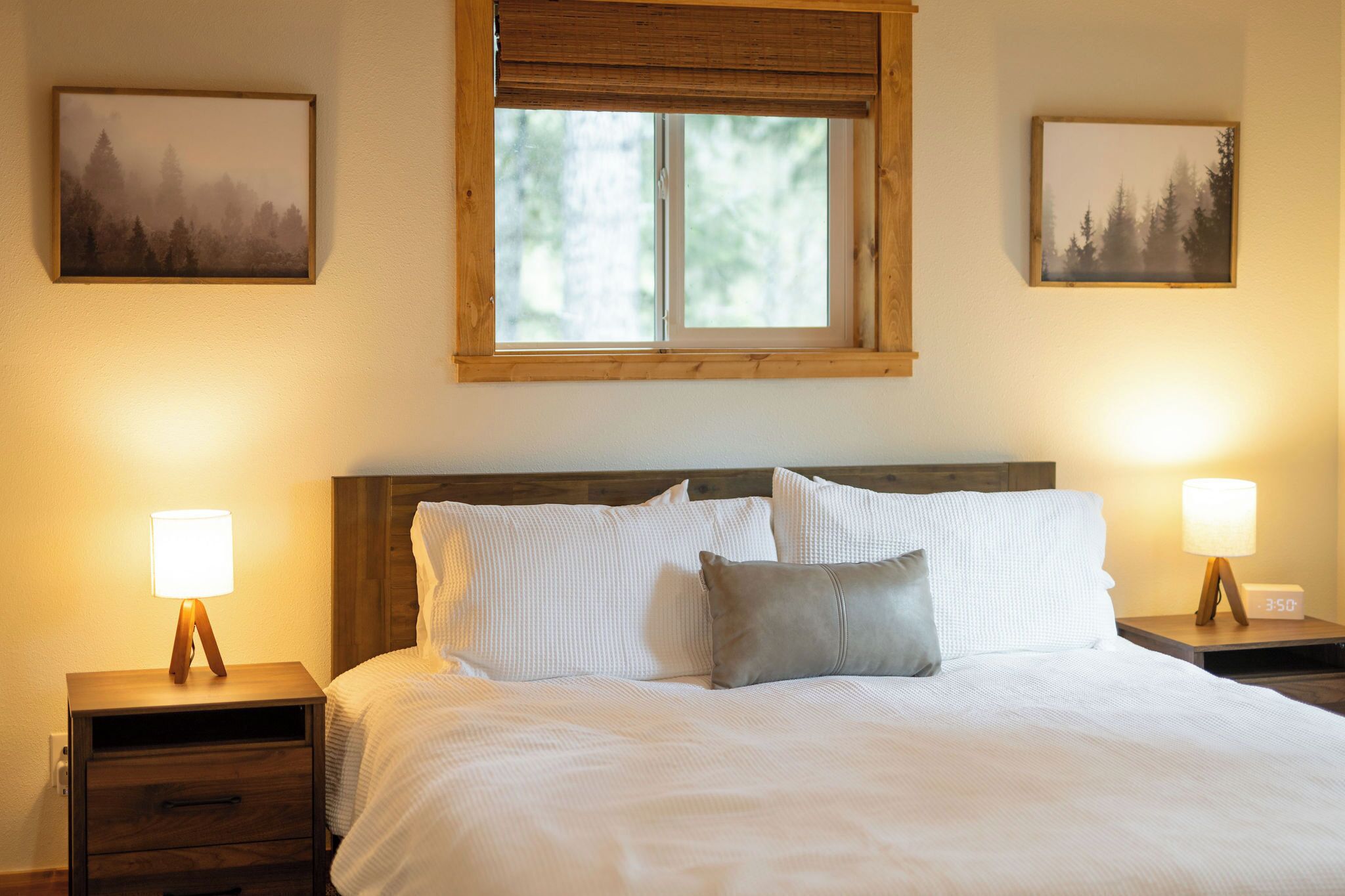 A bedroom with a large bed, two bedside tables with lamps, a window with a wooden frame, and two framed pictures on the walls.