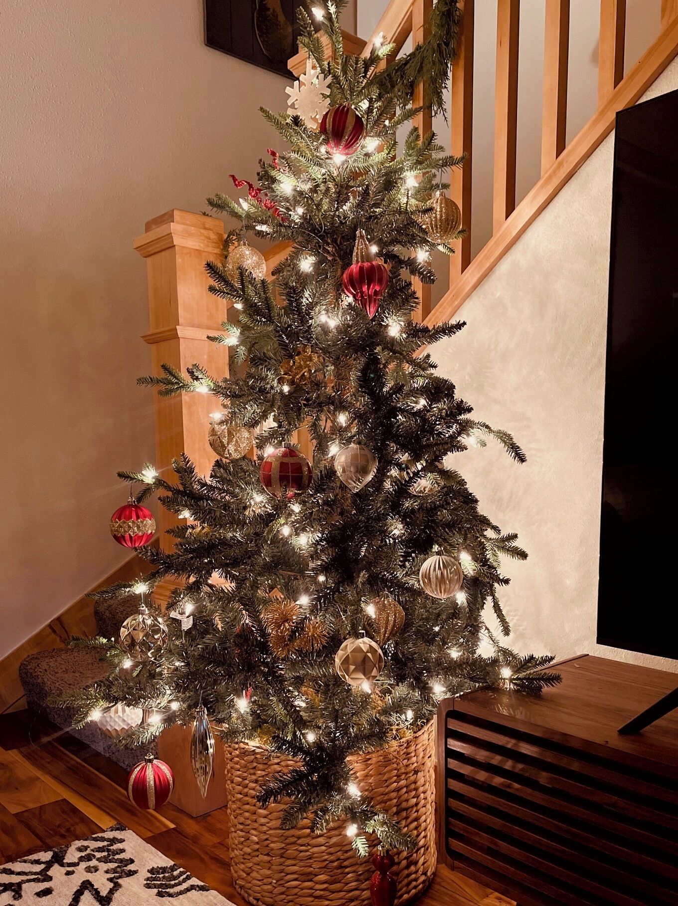 A Christmas tree with lights and ornaments, placed on a wooden floor near a staircase.